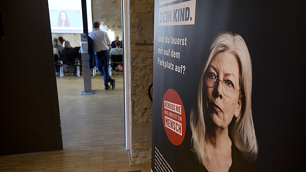 Foto eines Workshops, Blick durch die Tür in einen Raum mit Menschen, rechts steht ein Rollup mit Kampagnen-Design.