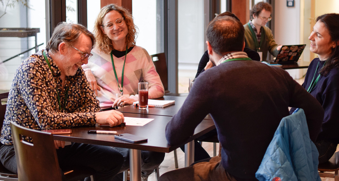Vier Menschen an einem Tisch, auf dem Tisch Stifte, Papiere und Getränke.