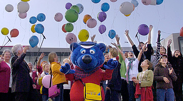Ansicht einer Menschengruppe, mit Luftballons und eine Figur im Kostüm von Käptn Blaubär