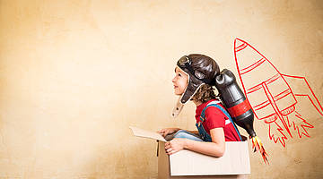 Kind in einem Karton sitzend, mit einer aus einer Öldose gebastelten Rakete auf dem Rücken