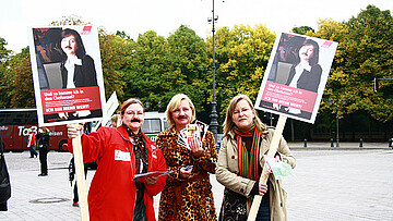 Drei Frauen halten Plakate und tragen aufgeklebte Schnauzbärte.