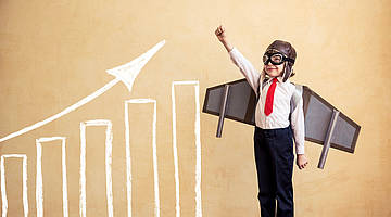 Boy with aeroplane wings attached in a superman pose in front of a chart with rising columns