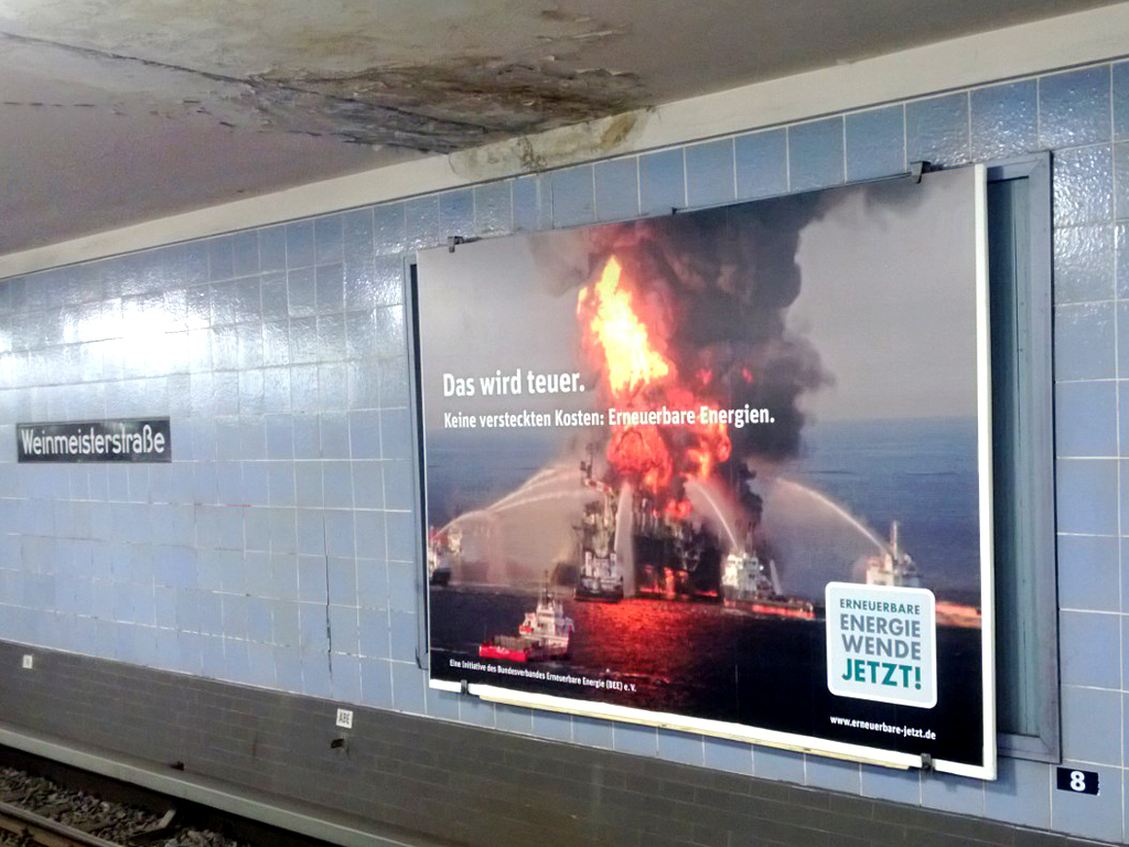 18-1_BEE_Weinmeister Bild einer brenenenden Ölplattform in einer U-Bahn Station. Über dem Poster ist ein schwarzer Fleck an der Decke