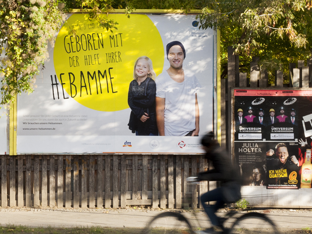 18-1 Lattenzaun big poster showing two kids "born with the help of their midwife" at the countryside
